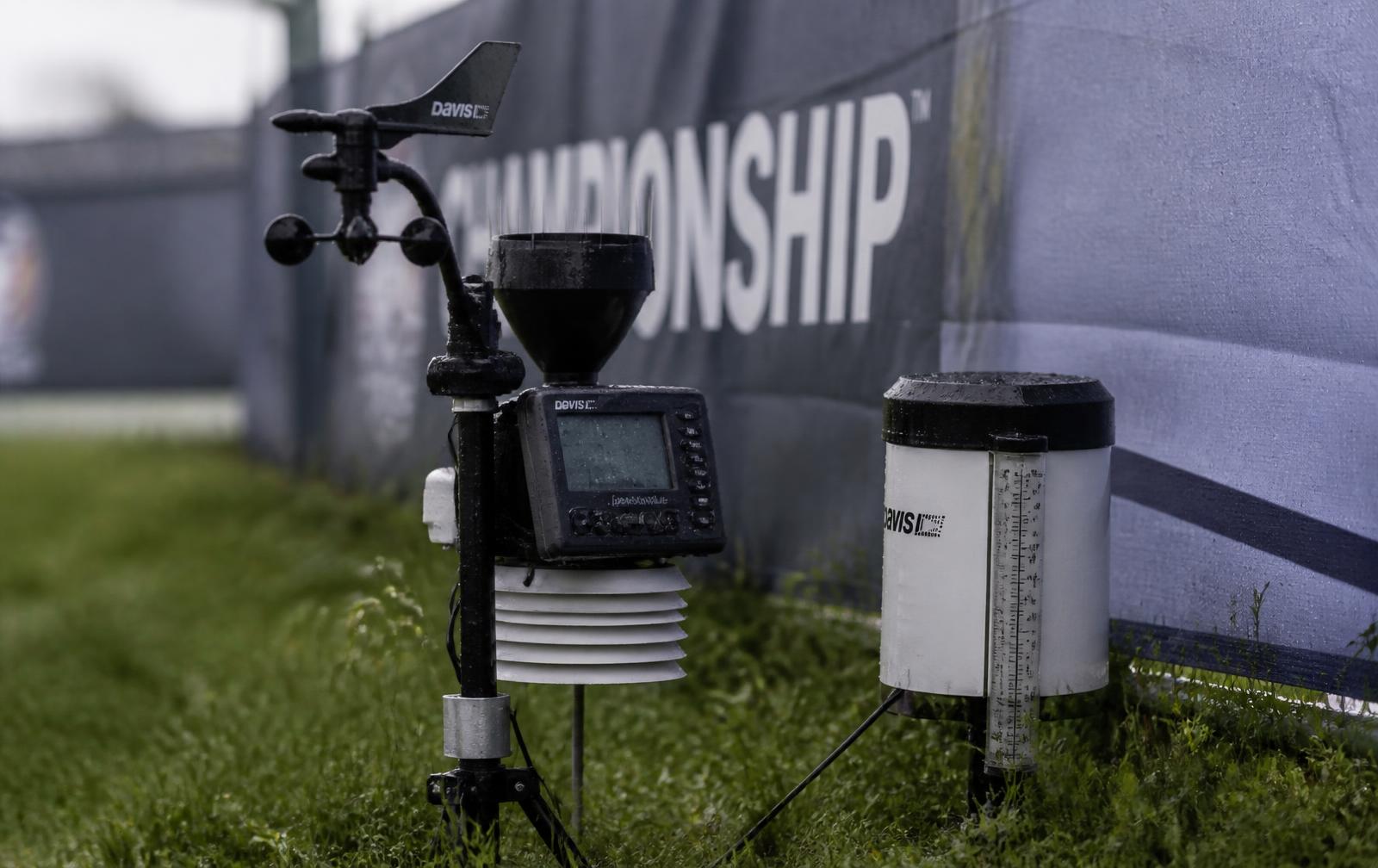 Close view of a weather monitoring instrument and rain gauge mounted at the edge of an open event field