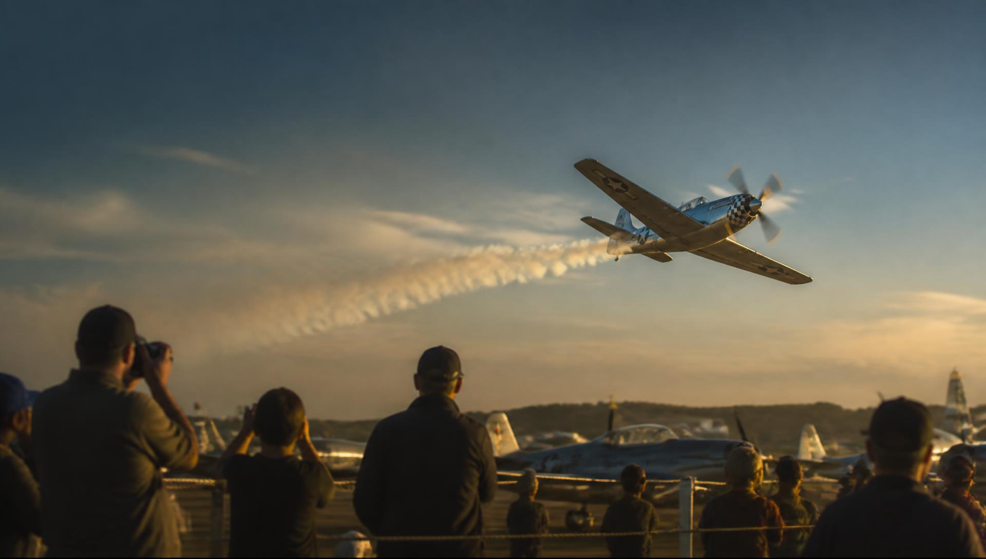 Spectators behind a barrier fence watching a line of parked propeller aircraft at a small airshow
