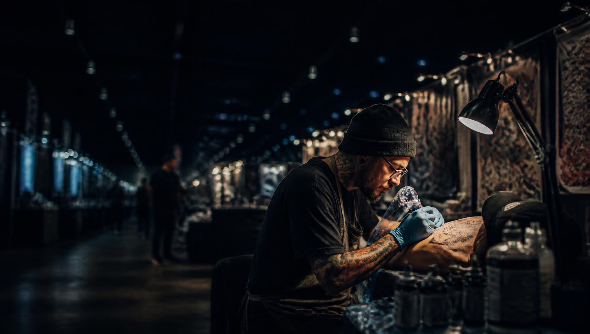 Tattoo artist in black gloves working on a client's forearm at a convention booth under an adjustable lamp