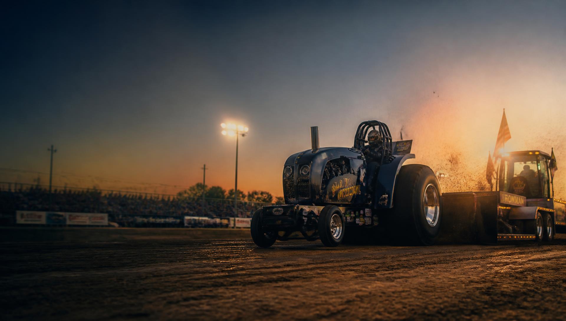 Modified orange tractor dragging a weight transfer sled down a dirt track under floodlights with a grandstand crowd behind barriers